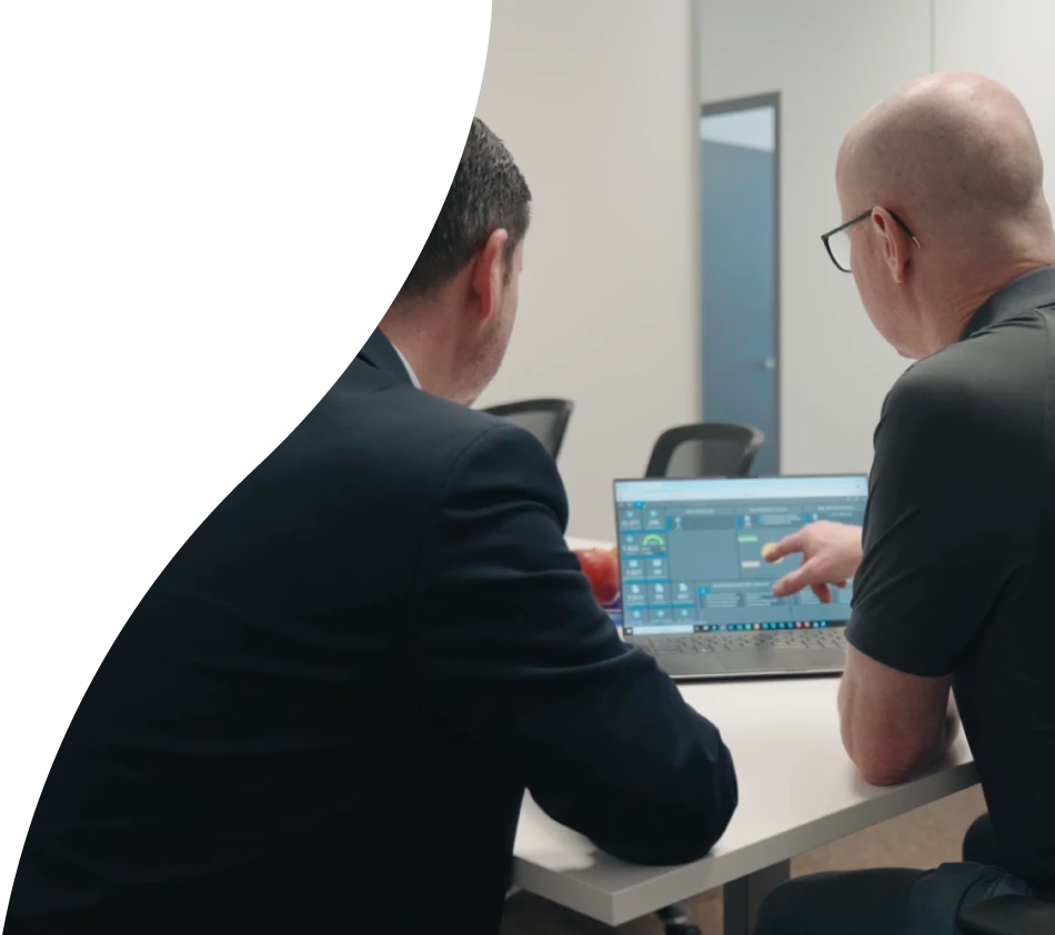 Two men are sitting with their backs to the camera in an office meeting room. On the table in front of them is a laptop with the crossbase dashboard open. The man on the right is pointing at the display and wearing glasses.