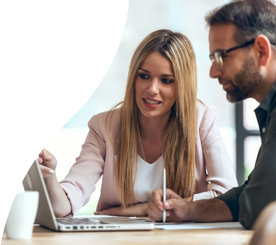 Zwei Personen, eine Frau und ein Mann, sitzen gemeinsam an einem Tisch und schauen konzentriert auf einen Laptop-Bildschirm. Die Frau zeigt auf den Bildschirm und erklärt etwas, während der Mann Notizen macht. Beide wirken in Ihre Arbeit oder eine Präsentation vertieft.