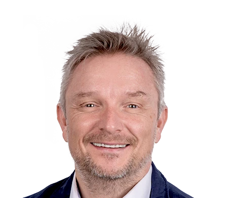 Holger Geck, Leiter Marketing und Produktdatenmanagement bei Gust. Alberts, lächelt in die Kamera. Er hat kurzes, graublondes Haar und einen kurzen Bart. Er trägt ein weißes Hemd und ein dunkles Sakko. Im Hintergrund ist eine geschwungene Wellenform in Weiß, Hellblau und Dunkelblau zu sehen.