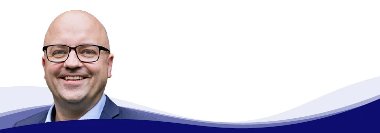 Tom Ruegenberg, Projektmanager bei Gebr. Kemper, lächelt und blickt in die Kamera. Er trägt eine Brille mit schwarzem Gestell, ein hellblaues Hemd mit offenem Kragen und ein graues Sakko darüber. Er hat eine Glatze und ist glatt rasiert.