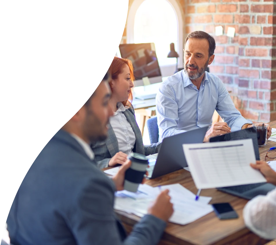 Four people in business attire are sitting at a wooden table in a modern office. They are engrossed in a conversation and working together with laptops, documents and coffee mugs. A window with daylight and a monitor can be seen in the background. The atmosphere is focused and professional.