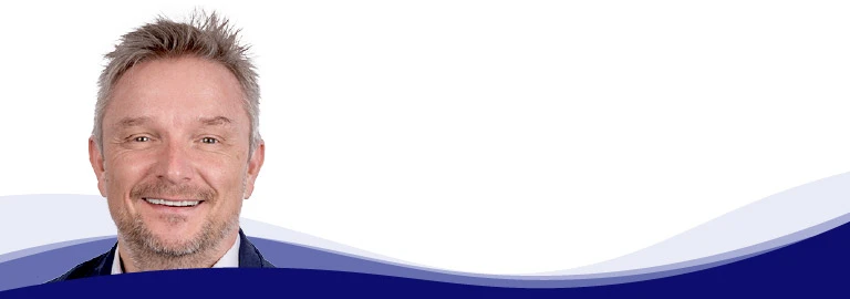 Holger Geck, directeur du marketing et de la gestion des données produits chez Gust. Alberts, sourit à la caméra. Il a les cheveux courts, blond grisâtre et une courte barbe. Il porte une chemise blanche et une veste sombre. En arrière-plan, on peut voir une forme de vague incurvée en blanc, bleu clair et bleu foncé.