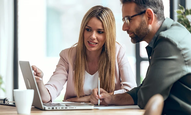 Deux personnes, une femme et un homme, sont assises ensemble à une table et regardent avec concentration l'écran d'un ordinateur portable. La femme montre l'écran et explique quelque chose pendant que l'homme prend des notes. Les deux semblent plongés dans leur travail ou dans une présentation.