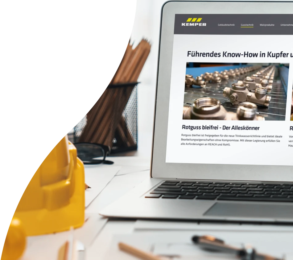 Un ordinateur portable ouvert sur une table montre l'écran de la boutique en ligne de KEMPER, qui a été réalisée avec crossbase. Sur la table, il y a des plans enroulés, un casque de protection pour les chantiers, une plante, des lunettes et un pot à crayons.