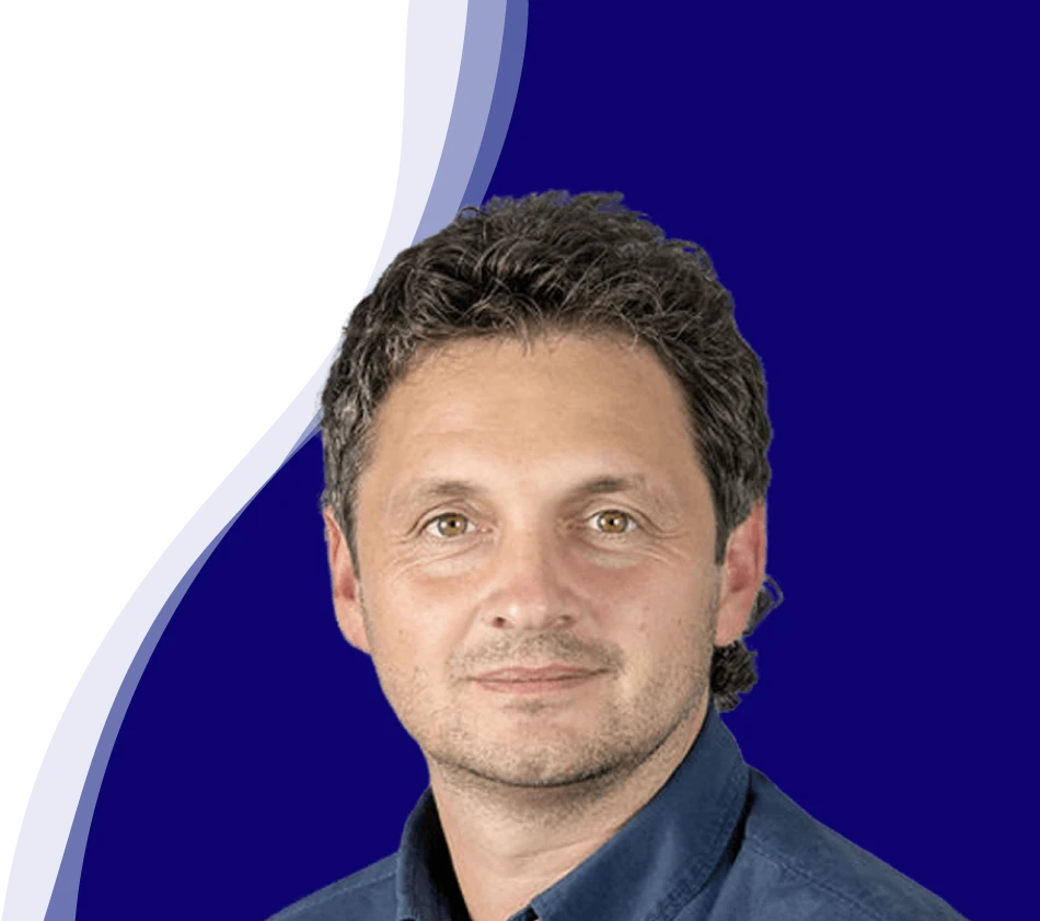 Reinhard Sutterlüty de l'entreprise meusburger sourit à la caméra. Il a les cheveux bouclés et foncés et porte une chemise bleu foncé. L'arrière-plan montre une forme de vague moderne en blanc, bleu clair et bleu foncé, qui s'étend en courbe du bas à gauche au haut à droite de l'image.