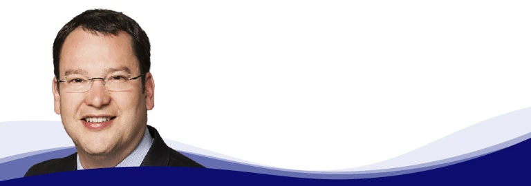 Dirk Amann, CTO Managing Director chez i.safe MOBILE, regarde la caméra en souriant. Il porte des lunettes sans monture, une chemise bleu clair et une veste sombre. Il a les cheveux courts, châtain foncé et un teint naturel.